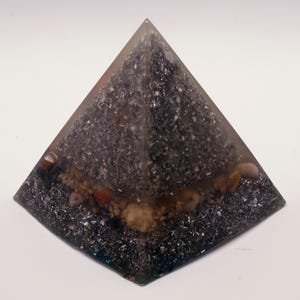 May include: A clear resin pyramid with a dark, speckled interior. The pyramid contains small stones and metallic flakes, creating a textured, layered appearance. The base is a dark, square shape.