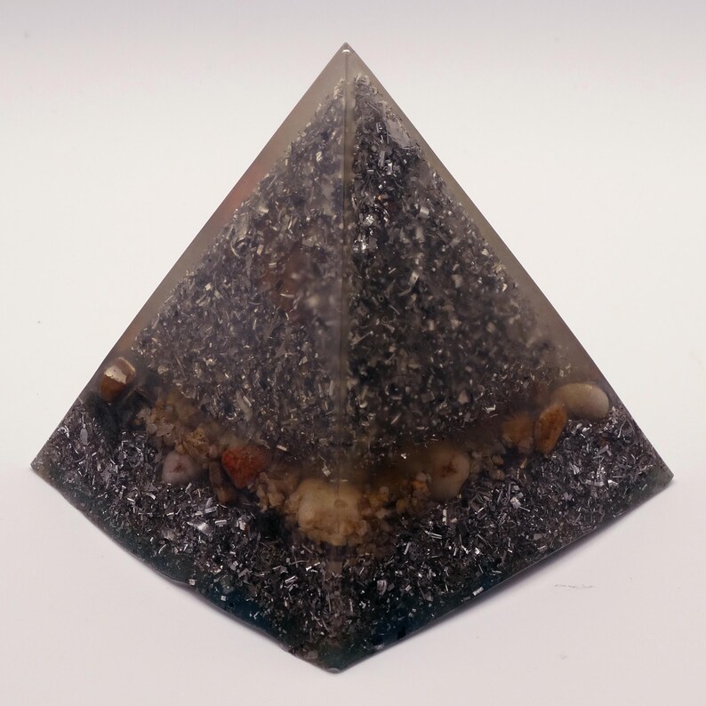 May include: A clear resin pyramid with a dark, speckled interior. The pyramid contains small stones and metallic flakes, creating a textured, layered appearance. The base is a dark, square shape.