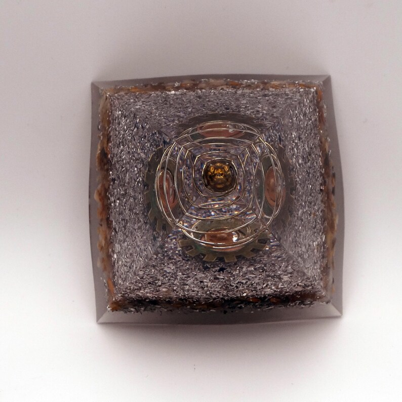 May include: A square pyramid object with a clear resin exterior. The interior has a silver granular material and a central metallic spiral design with a golden centre. The edges have a brown, textured appearance. The object is approximately 7 cm square.