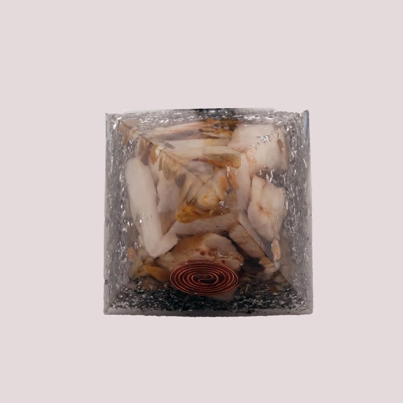 May include: A clear resin cube containing light-colored stones and a copper coil. The cube has a silver glitter-like appearance on the outside. The stones have a mix of white, beige, and brown hues.