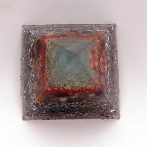 May include: A square orgone pyramid with a silver-colored exterior. The pyramid has a central turquoise-colored square, surrounded by a red border and copper coils. The sides are embedded with small stones.