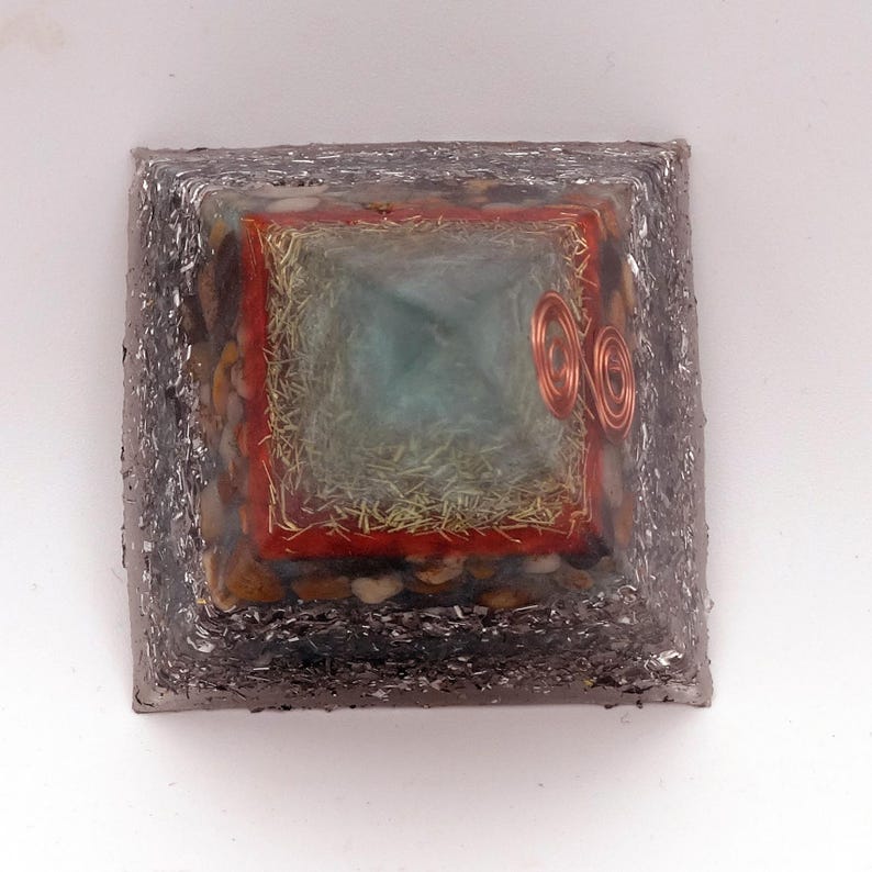 May include: A square orgone pyramid with a silver-colored exterior. The pyramid has a central turquoise-colored square, surrounded by a red border and copper coils. The sides are embedded with small stones.