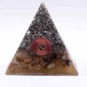 May include: A pyramid-shaped orgone energy generator. The pyramid is made of clear resin with embedded stones and metal fragments. A copper coil is centered on the front. The base is a light brown color.