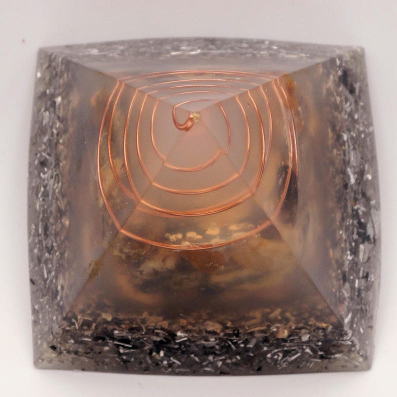May include: A square, clear resin pyramid with a copper coil at the center. The pyramid's base is a dark gray color with embedded metallic flakes. The interior has a light brown and beige pattern.
