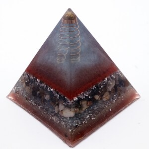 May include: A pyramid-shaped decorative object with a copper coil at the top. The pyramid has a red and gray gradient, with embedded stones and metallic flakes. The base is a darker red, transitioning to gray towards the top.