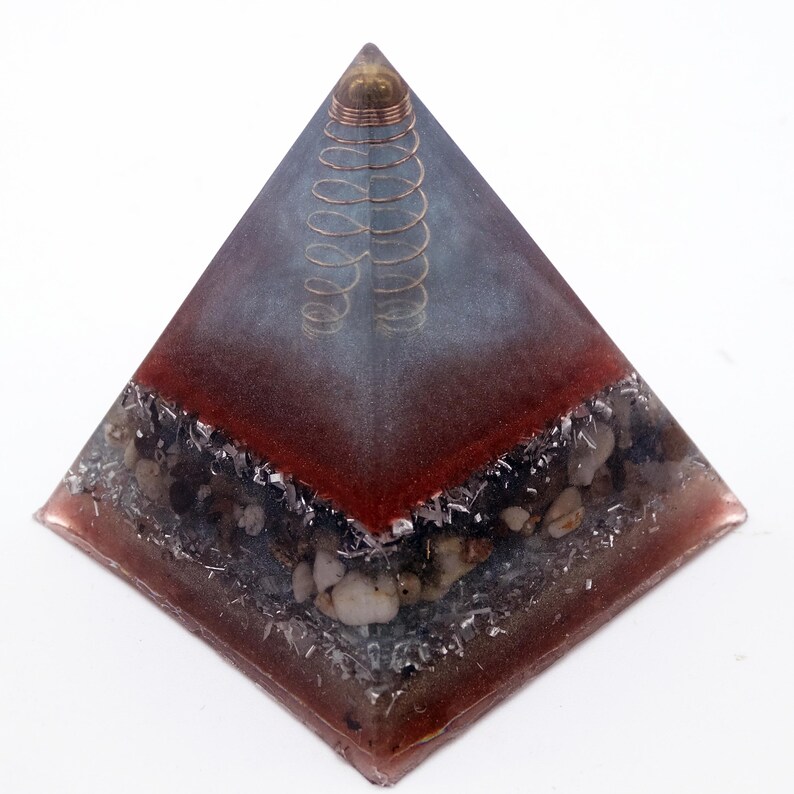 May include: A pyramid-shaped decorative object with a copper coil at the top. The pyramid has a red and gray gradient, with embedded stones and metallic flakes. The base is a darker red, transitioning to gray towards the top.