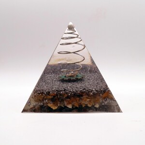 May include: A clear resin pyramid with a silver metal spiral and a small green gear. The base of the pyramid contains small, light-coloured stones and dark grey granules. A small, round, silver-coloured object sits at the top.