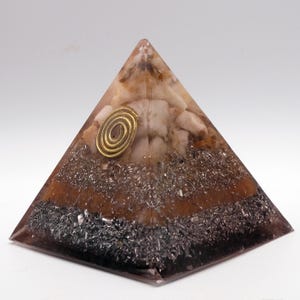 May include: A layered pyramid-shaped object with a clear, glossy exterior. The pyramid features a gold spiral accent and is filled with various materials, including brown, gray, and white elements. The base is dark, transitioning to lighter tones towards the top.