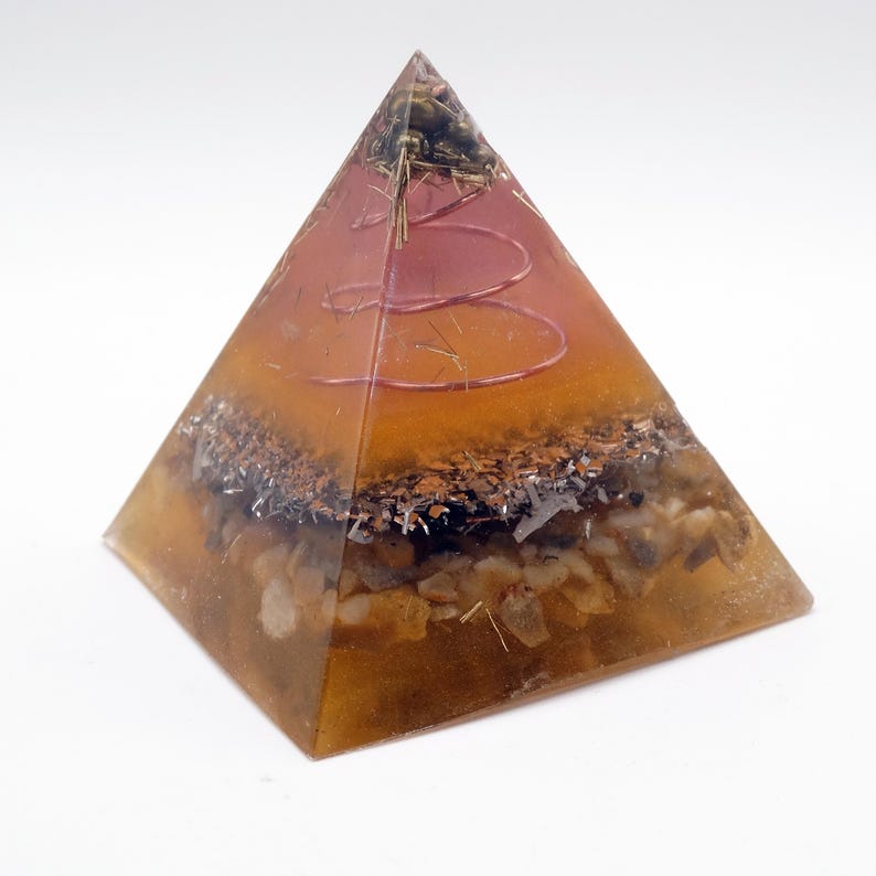 Peut inclure: Une pyramide transparente avec un design en couches. La base est de couleur brun dor&eacute;, avec une couche de petits cailloux et de particules sombres. Au-dessus, une spirale cuivr&eacute;e et de la mati&egrave;re v&eacute;g&eacute;tale sont visibles, enferm&eacute;es dans une r&eacute;sine ambr&eacute;e translucide.