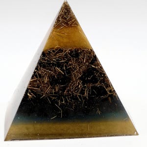 Puede incluir: Una pir&aacute;mide generadora de energ&iacute;a org&oacute;nica. La pir&aacute;mide est&aacute; hecha de resina transparente con capas de marr&oacute;n oscuro y dorado. Las capas contienen peque&ntilde;os trozos de metal dorado. La base de la pir&aacute;mide tiene un tono azul verdoso oscuro.