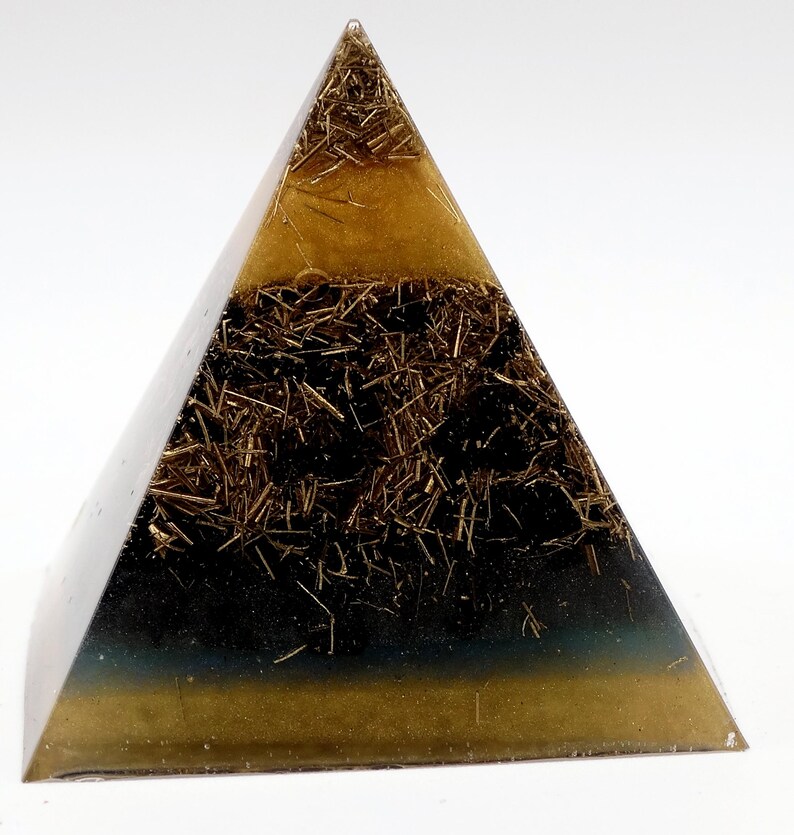 Puede incluir: Una pir&aacute;mide generadora de energ&iacute;a org&oacute;nica. La pir&aacute;mide est&aacute; hecha de resina transparente con capas de marr&oacute;n oscuro y dorado. Las capas contienen peque&ntilde;os trozos de metal dorado. La base de la pir&aacute;mide tiene un tono azul verdoso oscuro.