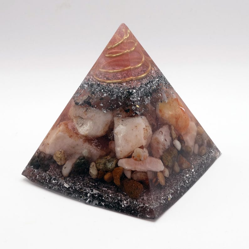 May include: A pyramid-shaped decorative object with a clear resin exterior. The pyramid contains various stones in shades of pink, white, and brown, along with a metallic spiral at the top. The base is a dark, speckled color.