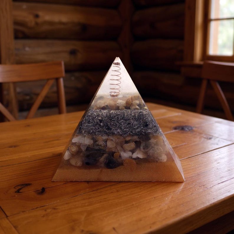 Puede incluir: Objeto decorativo en forma de pir&aacute;mide, con capas visibles de arena, piedras y un resorte en la parte superior, encapsulado en resina transparente. Colocado sobre una mesa de madera.