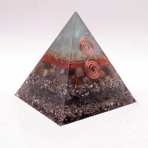 May include: A layered pyramid sculpture with a clear apex and a copper wire spiral. The pyramid's layers include red, brown, and silver materials. The base is a dark, textured material. The pyramid is a decorative item.