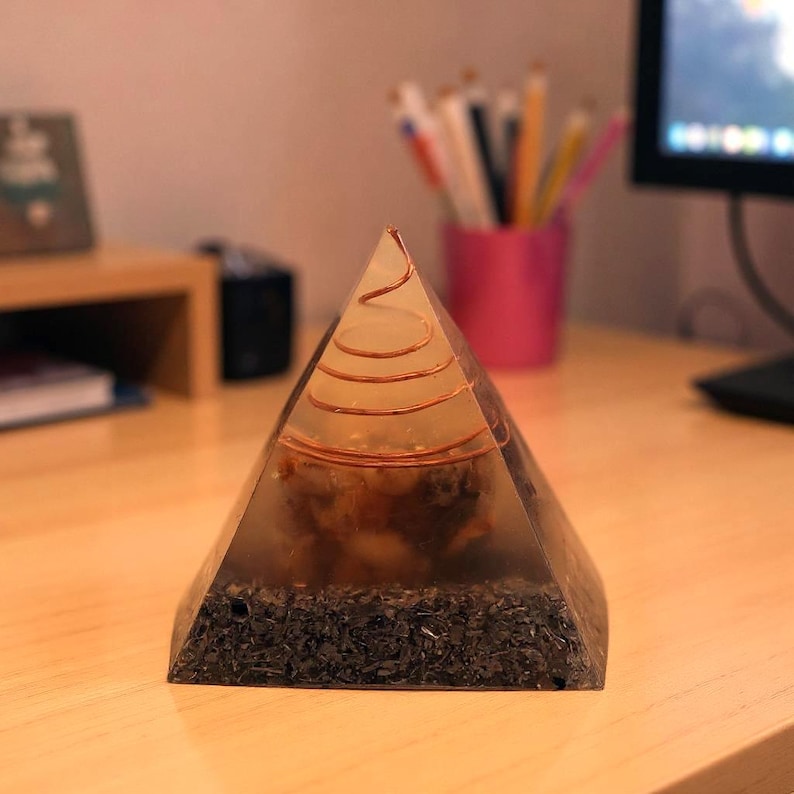 May include: A clear pyramid-shaped orgone energy generator with a copper coil at the top. The pyramid contains dark, organic material and is set on a wooden surface. The pyramid is a decorative item.