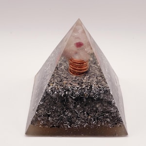 May include: A clear pyramid-shaped orgone energy generator. The pyramid contains a copper coil, a pink crystal, and dark gray metallic fragments. The pyramid is on a white background.