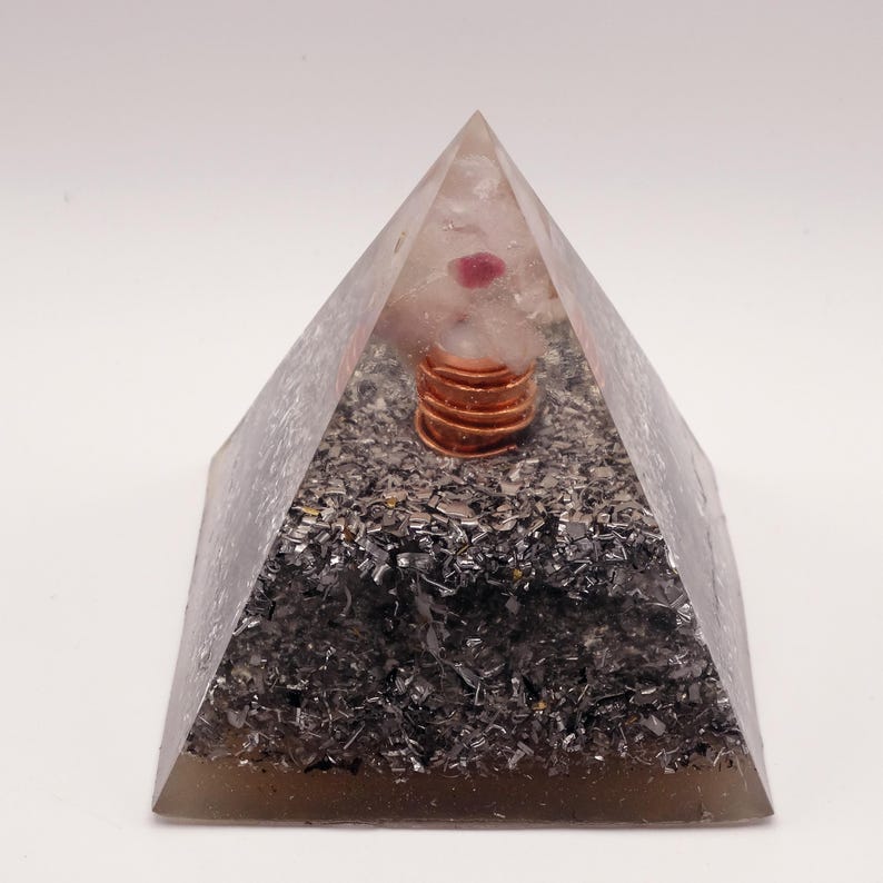 May include: A clear pyramid-shaped orgone energy generator. The pyramid contains a copper coil, a pink crystal, and dark gray metallic fragments. The pyramid is on a white background.