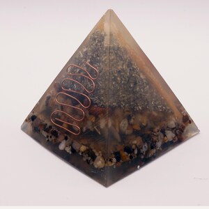 May include: A clear resin pyramid with embedded stones, crystals, and a copper coil. The pyramid has a layered appearance with a mix of colors, including brown, gray, and clear. The copper coil is a prominent feature, adding a decorative element.