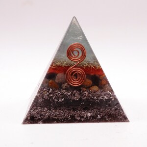 May include: A layered pyramid-shaped orgone energy generator. The pyramid features a clear resin exterior with copper wire spirals and various embedded materials, including red, brown, and black elements. The pyramid is designed to be a decorative and spiritual item.