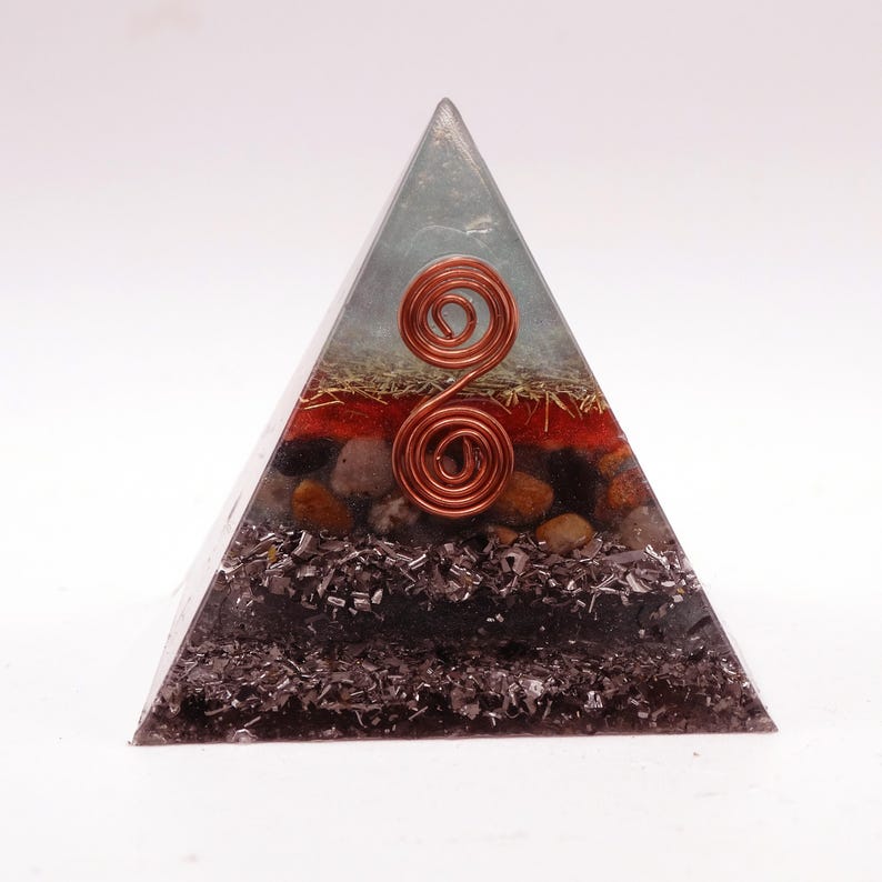 May include: A layered pyramid-shaped orgone energy generator. The pyramid features a clear resin exterior with copper wire spirals and various embedded materials, including red, brown, and black elements. The pyramid is designed to be a decorative and spiritual item.
