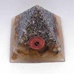 May include: A pyramid-shaped orgone energy generator. The top section is filled with a dark, metallic material, while the base has a tan and brown appearance. A copper coil is embedded on the front. The pyramid is made of resin.