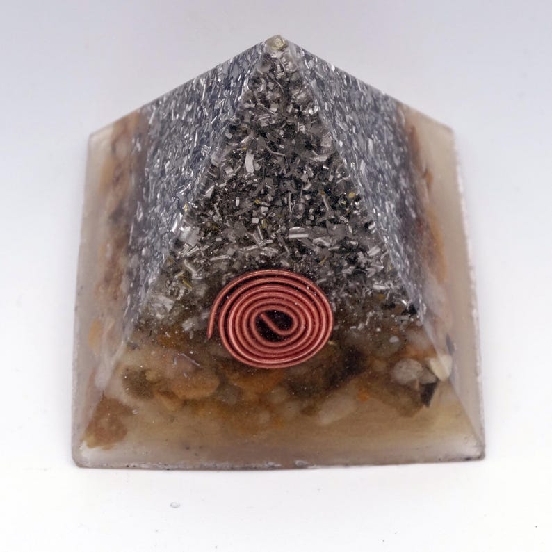 May include: A pyramid-shaped orgone energy generator. The top section is filled with a dark, metallic material, while the base has a tan and brown appearance. A copper coil is embedded on the front. The pyramid is made of resin.