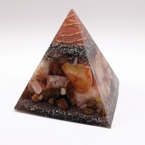 May include: A pyramid-shaped orgone energy generator. The pyramid is made of clear resin, with layers of stones, crystals, and a copper coil at the top. The stones are various colors, including orange, white, and brown. The pyramid sits on a white surface.