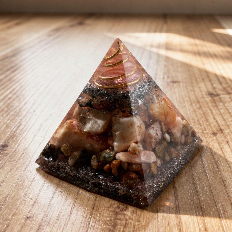 May include: A clear pyramid-shaped object with embedded stones and a gold spiral at the top. The pyramid contains various colors of stones, including beige, brown, and green. The pyramid is sitting on a wooden surface.