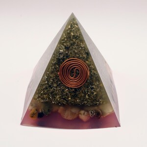 May include: A clear pyramid-shaped orgone energy generator. The pyramid has a copper coil at the center and is filled with green and brown organic material. The base is a translucent pink color, with small stones visible.
