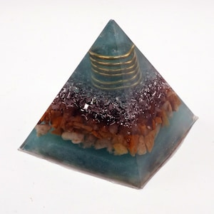 May include: A teal pyramid ornament with a gold coil at the apex. The pyramid is filled with a mix of brown, black, and white stones. The pyramid is made of a translucent material.