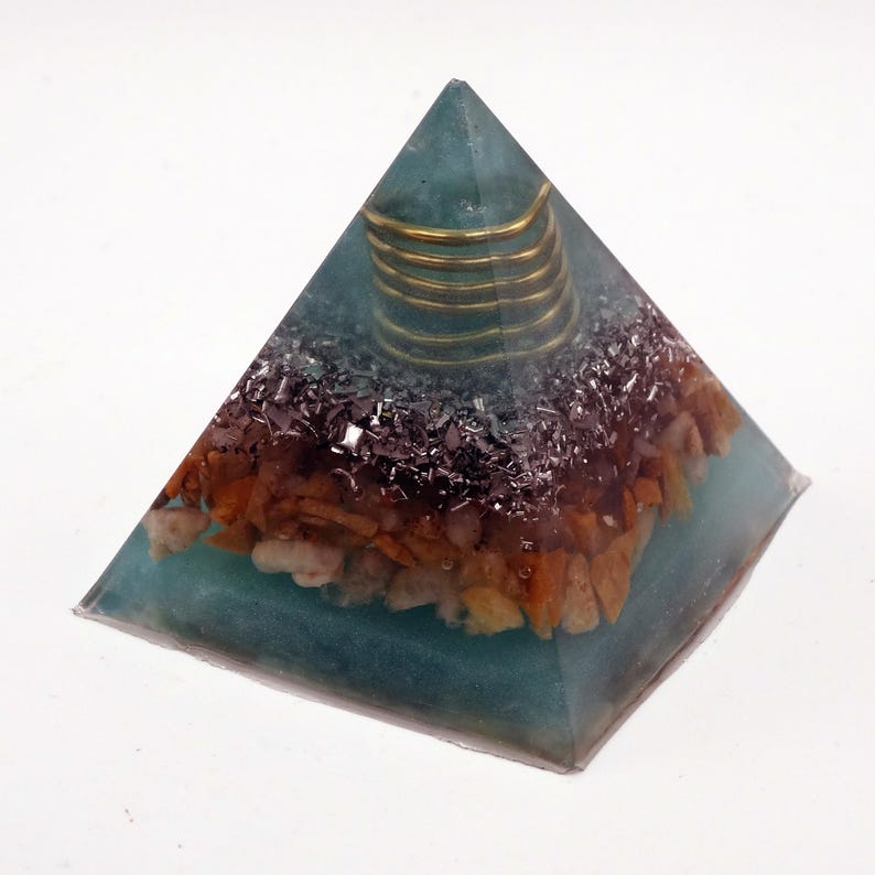 May include: A teal pyramid ornament with a gold coil at the apex. The pyramid is filled with a mix of brown, black, and white stones. The pyramid is made of a translucent material.