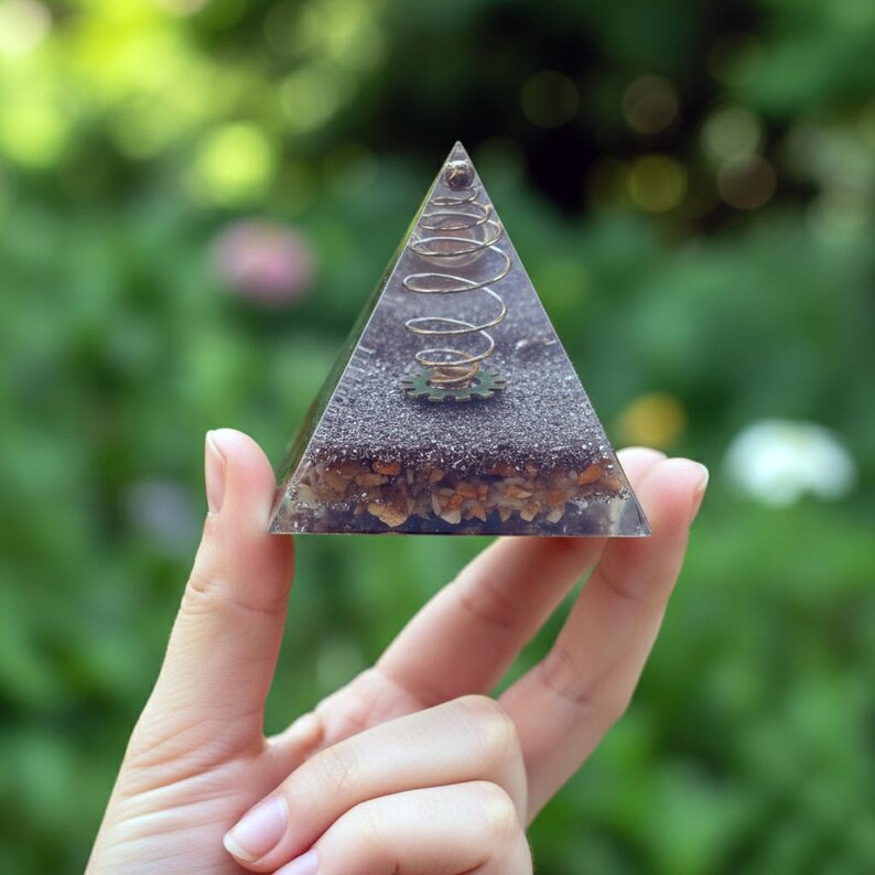 May include: A clear resin pyramid with embedded decorative elements. Inside, a copper-coloured coil and a small gear are visible, along with layers of sand and small stones. The pyramid is held against a blurred green background.
