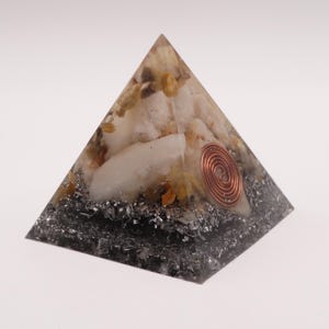 May include: A pyramid-shaped orgone energy generator. The pyramid features a copper coil, quartz crystals, and a dark gray base. The top section has white and yellow crystals. The pyramid is on a white background.