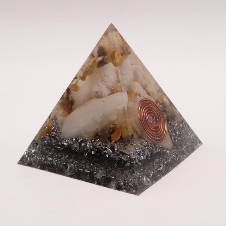 May include: A pyramid-shaped orgone energy generator. The pyramid features a copper coil, quartz crystals, and a dark gray base. The top section has white and yellow crystals. The pyramid is on a white background.