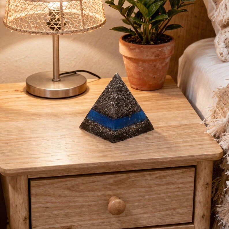 Peut inclure: Objet d&eacute;coratif en forme de pyramide noire et grise avec une bande bleue. La pyramide est pos&eacute;e sur une table de chevet en bois, &agrave; c&ocirc;t&eacute; d'une lampe et d'une plante en pot. La pyramide mesure environ 7,6 cm de haut.