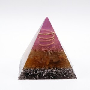 May include: A pyramid-shaped orgone energy generator. The top is a translucent purple, with a copper coil and a layer of pink and brown material. The middle section is amber and the base is black. This decorative piece is designed to promote positive energy.