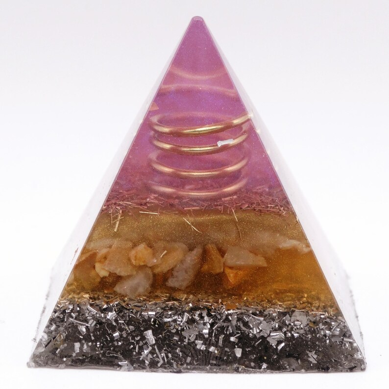 May include: A clear pyramid-shaped resin sculpture with a layered design. The top layer is a pinkish-purple hue with a gold coil, followed by a gold layer, then a layer of orange-toned stones, and a bottom layer of dark gray metallic flakes.