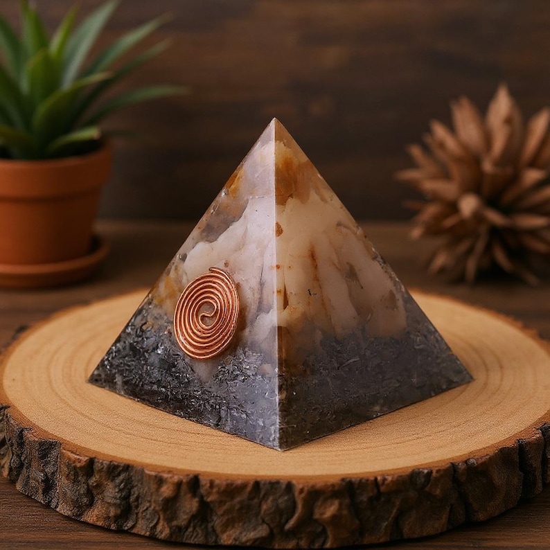 May include: A handcrafted pyramid-shaped orgone energy generator. The pyramid features a copper spiral and a mix of white, brown, and gray tones. It sits on a wooden slice, with a small potted plant in the background.