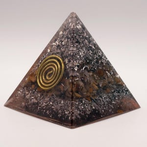 May include: A pyramid-shaped orgone energy generator. The pyramid is a translucent brown with embedded silver-colored metal shavings and small stones. A gold-colored spiral is affixed to the front of the pyramid.