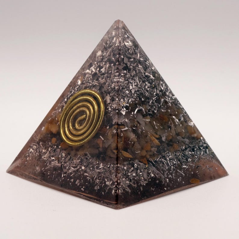 May include: A pyramid-shaped orgone energy generator. The pyramid is a translucent brown with embedded silver-colored metal shavings and small stones. A gold-colored spiral is affixed to the front of the pyramid.