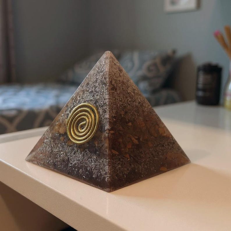 May include: A brown pyramid-shaped orgone energy generator with a gold-colored spiral design on the front. The pyramid is made of a translucent material with a speckled appearance. The pyramid sits on a white surface.