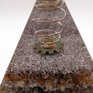 May include: A clear resin pyramid with embedded metallic flakes and small stones. A brass-coloured spring and a gear are set within the pyramid. The pyramid has a steampunk or industrial design.