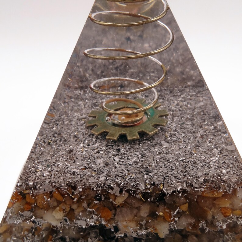 May include: A clear resin pyramid with embedded metallic flakes and small stones. A brass-coloured spring and a gear are set within the pyramid. The pyramid has a steampunk or industrial design.