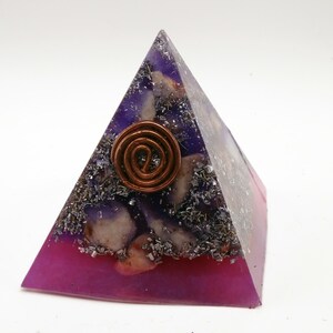 May include: A pyramid-shaped orgone energy generator with a copper coil at the top. The pyramid is made of purple and pink resin, with embedded crystals and metal shavings. The pyramid is on a white background.