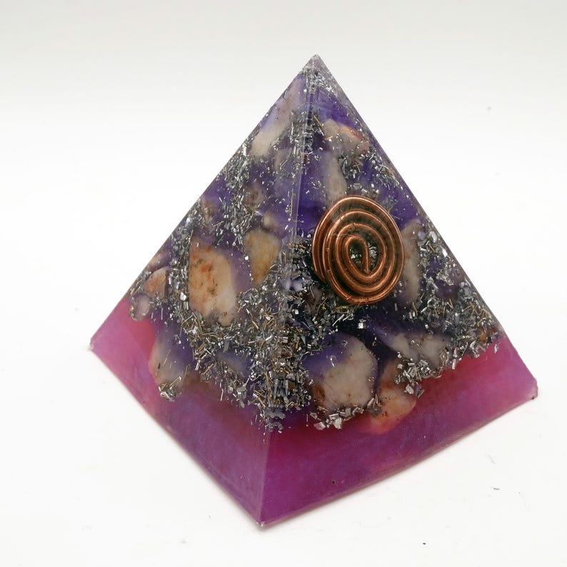 May include: A purple and pink pyramid-shaped orgone energy generator. The pyramid features a copper coil at the top, with a mix of purple resin, silver flakes, and beige stones. The base is a solid, vibrant pink.