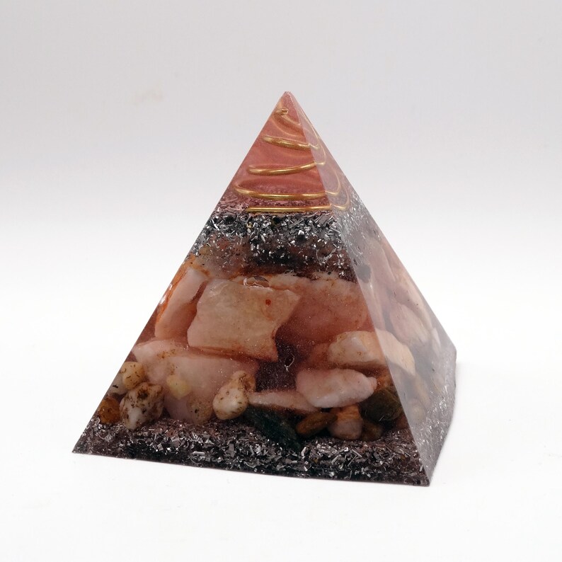 May include: A pyramid-shaped decorative object with a clear resin exterior. The interior features a mix of pink and white stones, dark gray metallic flakes, and gold-colored wire at the apex. The pyramid is set against a white background.
