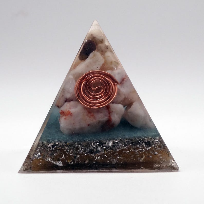 May include: A triangular orgone pyramid with a copper coil at the center. The pyramid is filled with white and beige stones, blue resin, and dark gray metallic flakes. The pyramid is on a white background.