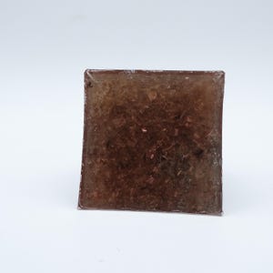 May include: A square, translucent block with a brown, textured interior. The block appears to be made of resin or a similar material, with small, reflective particles embedded throughout. The edges are slightly rounded, and the overall appearance is smooth.