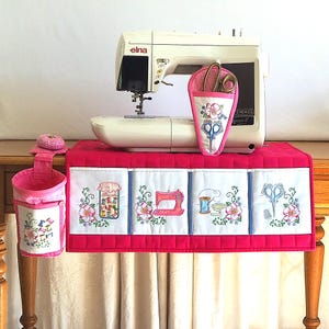 May include: A white Elna sewing machine with a pink cover and accessories. The cover has embroidered panels with sewing-themed designs. A pink pincushion, scissor holder, and a small bin are also visible.