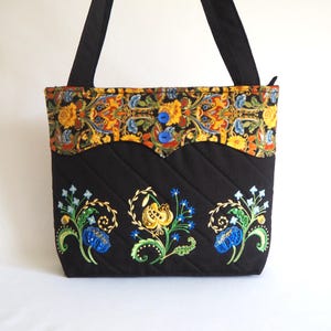 May include: A black quilted tote bag featuring two blue buttons and a floral patterned upper band. The lower section displays three embroidered floral motifs in blue, yellow, and green. The bag includes two black shoulder straps for carrying.